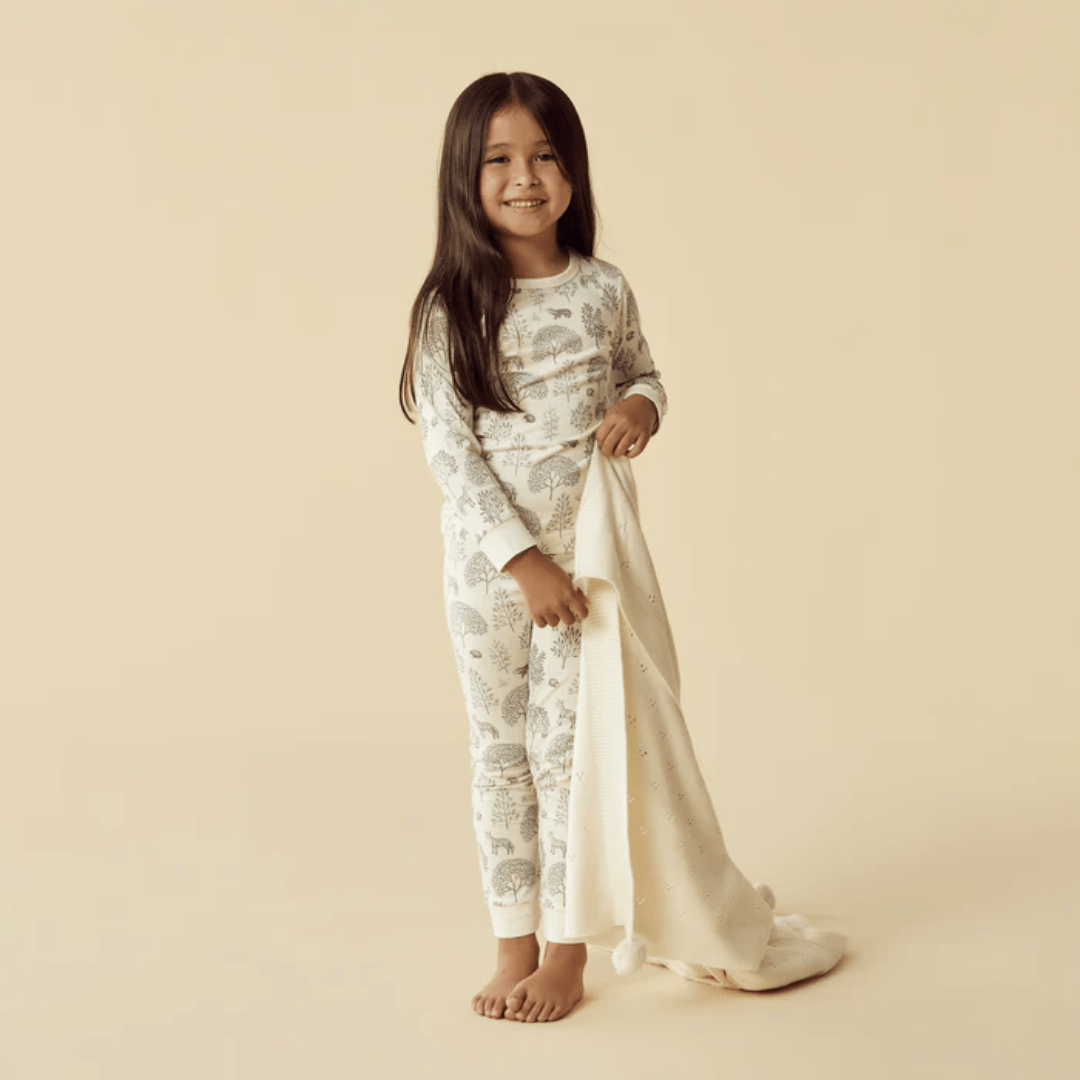 A young child wearing Wilson & Frenchy Organic Long Sleeved Pyjamas stands barefoot on a beige background, holding a light-colored blanket and smiling.