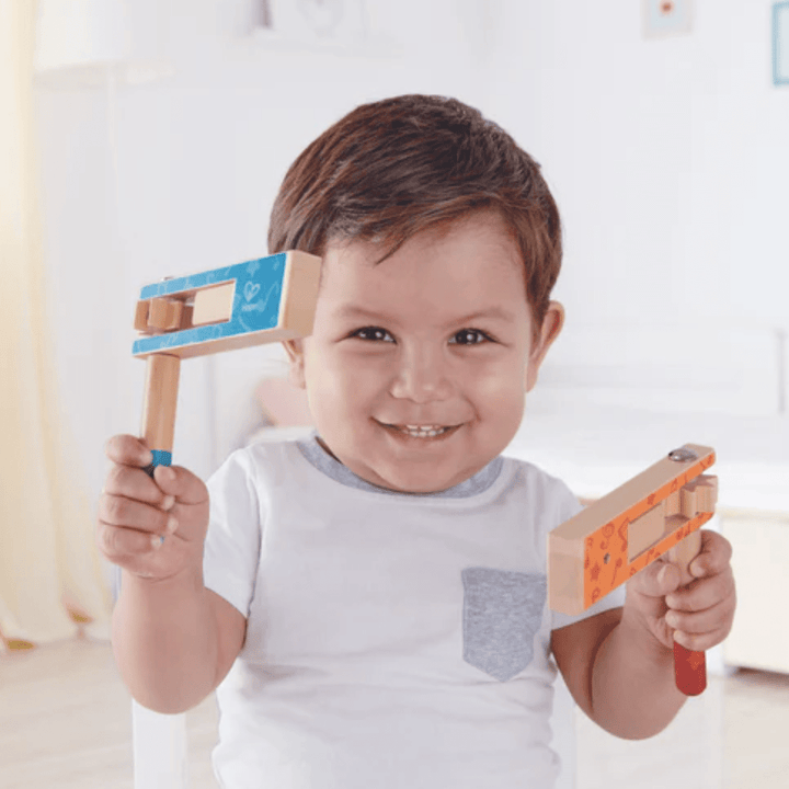 A toddler smiling and shaking the Hape Cheer a Long Noisemaker - Lucky Last in blue with both hands, demonstrating excellent hand-eye coordination while standing indoors.