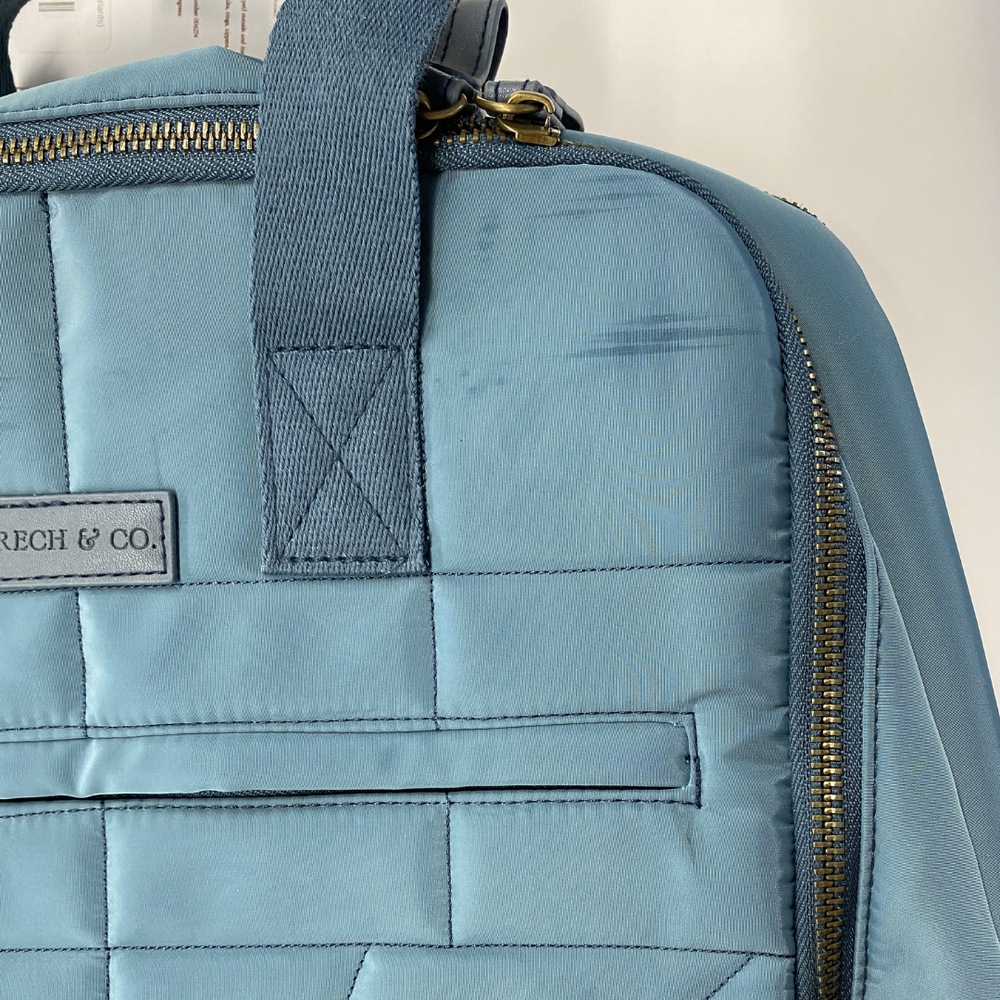 Close-up of a light blue, quilted fabric bag with a zipper and strap. A patch on the bag reads "GRECH & CO." This chic outlet item is perfect for those looking for eco-friendly backpacks. The background is white.