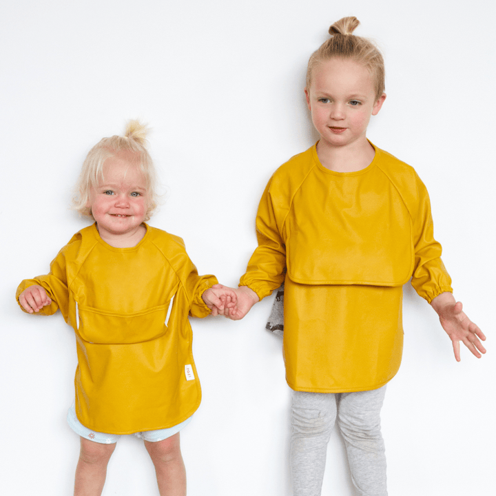 Two toddlers stand against a white background, both wearing yellow long-sleeve shirts. The older child on the right is also wearing light grey leggings, while the younger child wears light blue shorts and a colorful Zazi Recycled Full-Sleeved Bib from Zazi that is waterproof and stain-resistant, keeping their outfit clean.