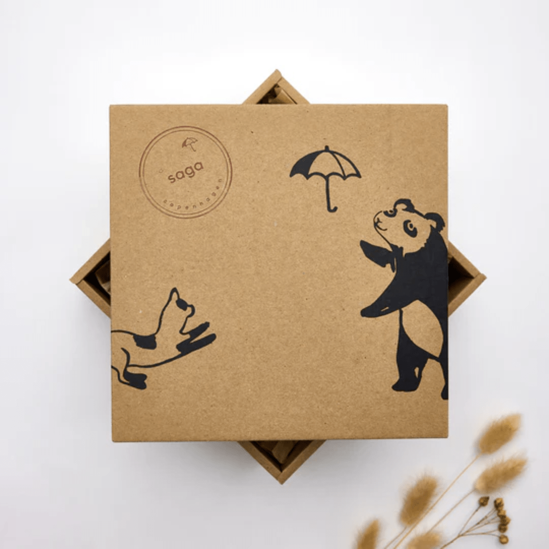 A brown square box features black illustrations of a panda holding an umbrella and a dog. The Saga Copenhagen branding is highlighted by a round stamp with "saga" in the top left corner. Its eco-friendly packaging details are complemented by visible dried flowers in the bottom right, marking it as the Saga Copenhagen Gift Box - Elina.