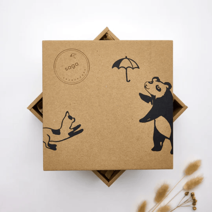 A brown square box features black illustrations of a panda holding an umbrella and a dog. The Saga Copenhagen branding is highlighted by a round stamp with "saga" in the top left corner. Its eco-friendly packaging details are complemented by visible dried flowers in the bottom right, marking it as the Saga Copenhagen Gift Box - Elina.