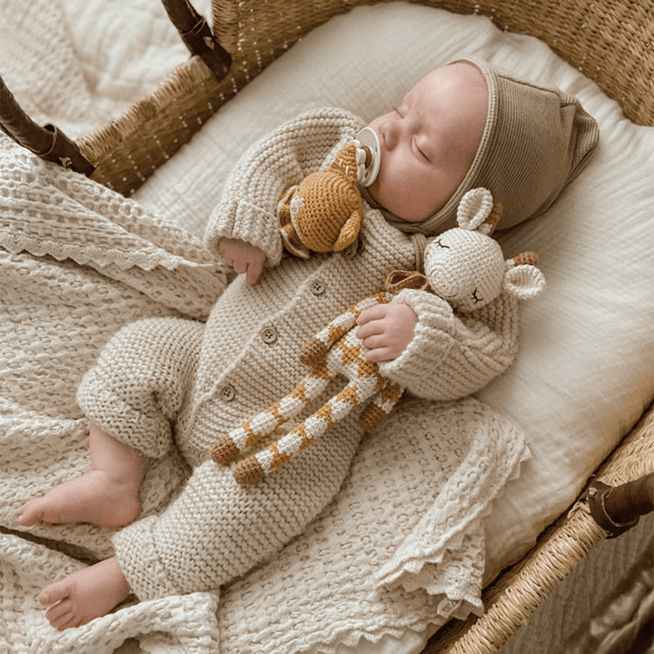 A baby in knitted clothing sleeps in a wicker crib, clutching a Patti Oslo Organic Cotton Goldie Giraffe and pacifier, on a cream-colored blanket.