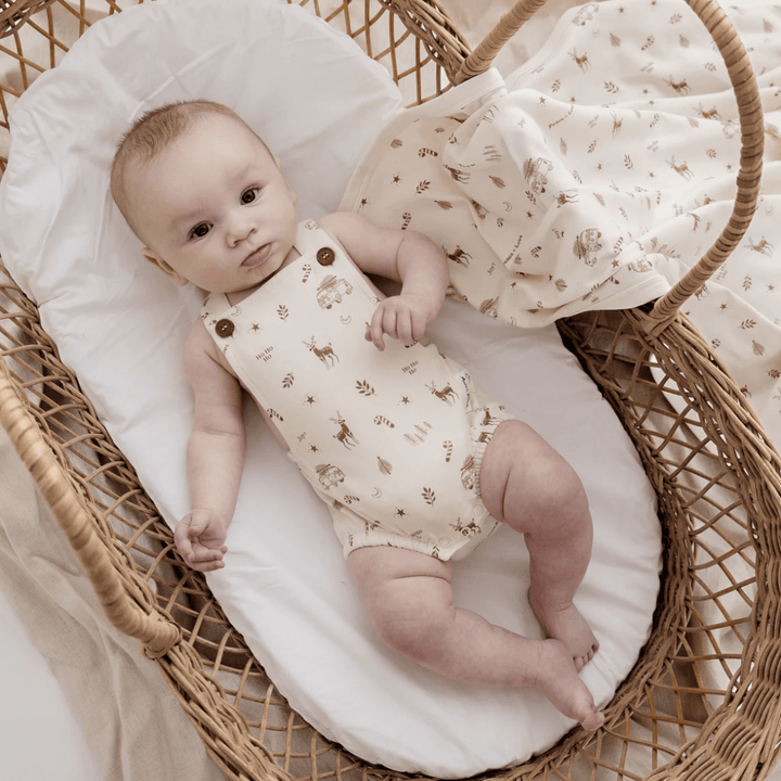 A baby rests in a wicker bassinet dressed in the Aster & Oak Organic Happy Holidays Playsuit, crafted from GOTS-certified cotton, showcasing a cream color with a nature print. A matching blanket is elegantly placed nearby.