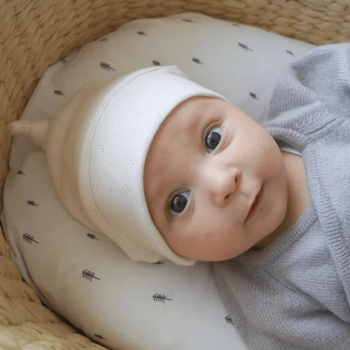 A baby nestled in a basket, sporting the cozy Lola & Me Organic Top Knot Beanie, surrounded by handmade baby accessories.