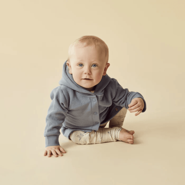 A baby wearing a blue Wilson & Frenchy Organic Quilted Jacket and patterned pants sits on a beige background, looking forward.