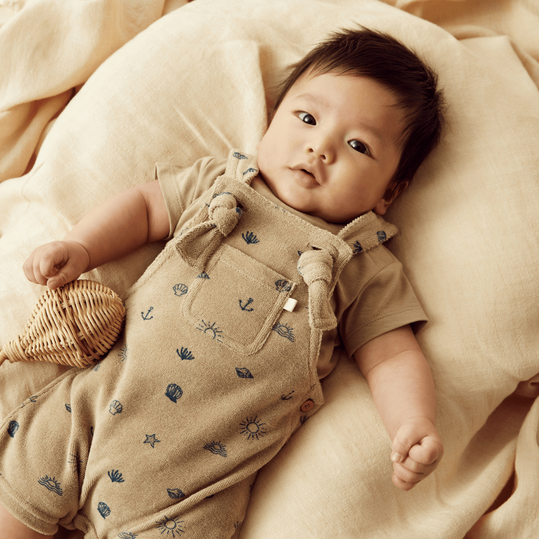 A baby is laying on a bed with a stuffed animal, dressed in Wilson & Frenchy Organic Terry Overalls - LUCKY LAST - MOSS - 3-6 MONTHS ONLY with adjustable straps.