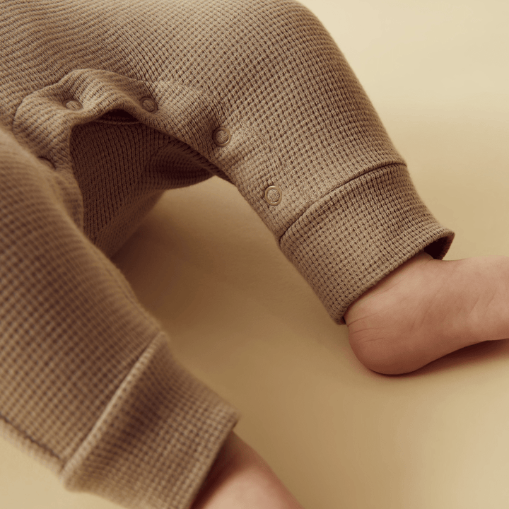 Close-up of a baby's legs and feet wearing Wilson & Frenchy Organic Waffle Overalls in Brilliant Blue, made from GOTS-certified organic cotton, set against a matching beige background.