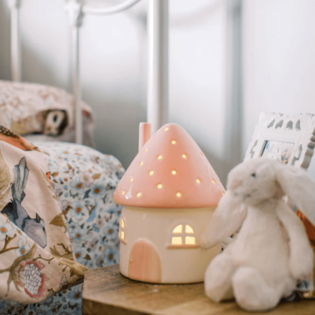 A cozy bedroom scene exudes whimsical charm with a plush bunny toy, a floral-patterned bedspread, and the Little Belle Nightlights Ceramic Nightlight – LUCKY LASTS – FAIRY HOUSE ONLY casting a soft glow in the background.