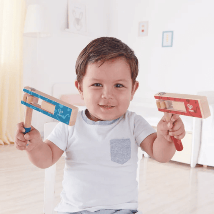 A toddler sits on a chair in a well-lit room, smiling and holding the Hape Cheer a Long Noisemaker in each hand, one blue and one red.