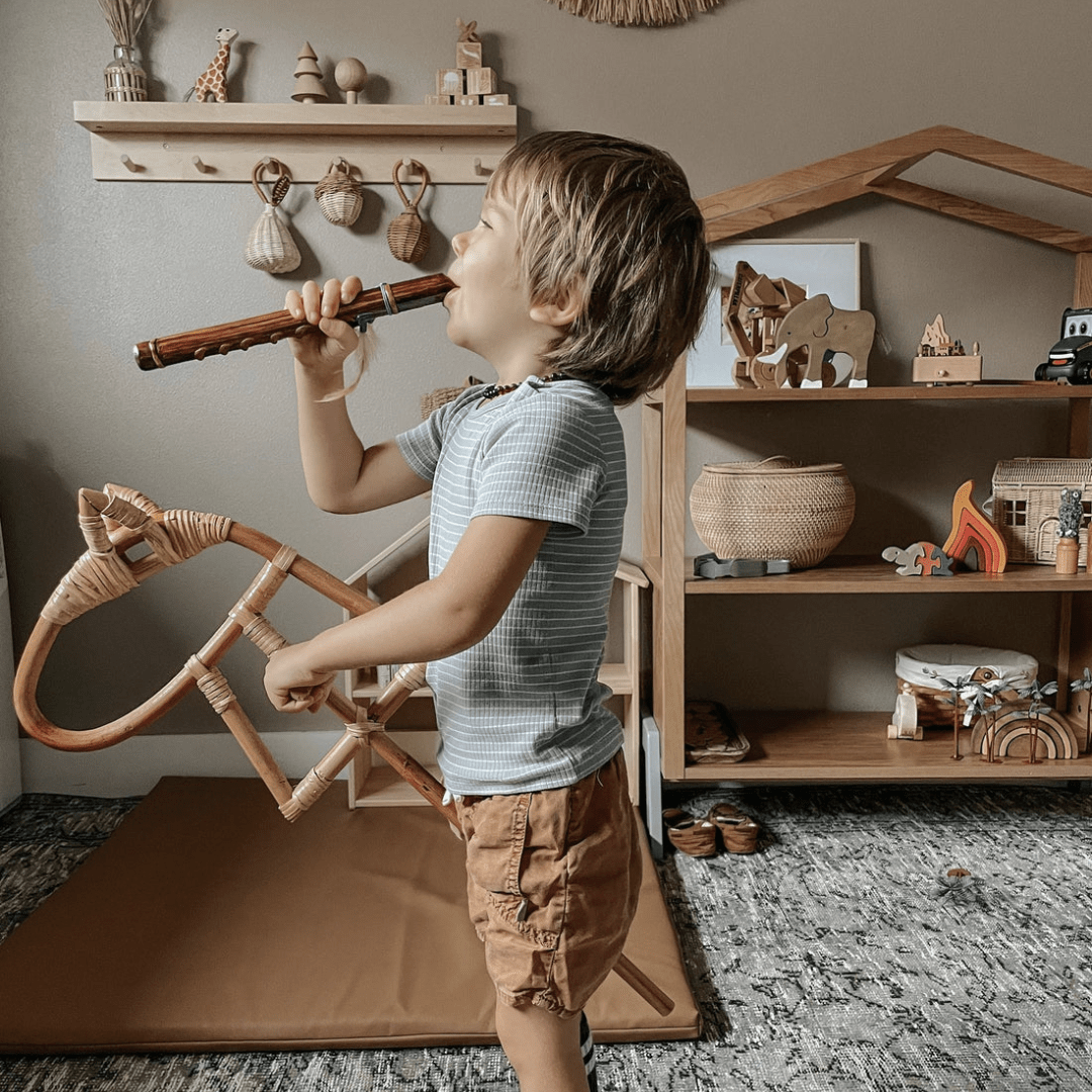 A child wearing a striped shirt and brown shorts plays a wooden recorder in a playroom filled with wooden toys, natural decor, and the Classical Child Rattan Hobby Horse - LUCKY LAST, fostering imaginative play while emphasizing eco-responsibility.