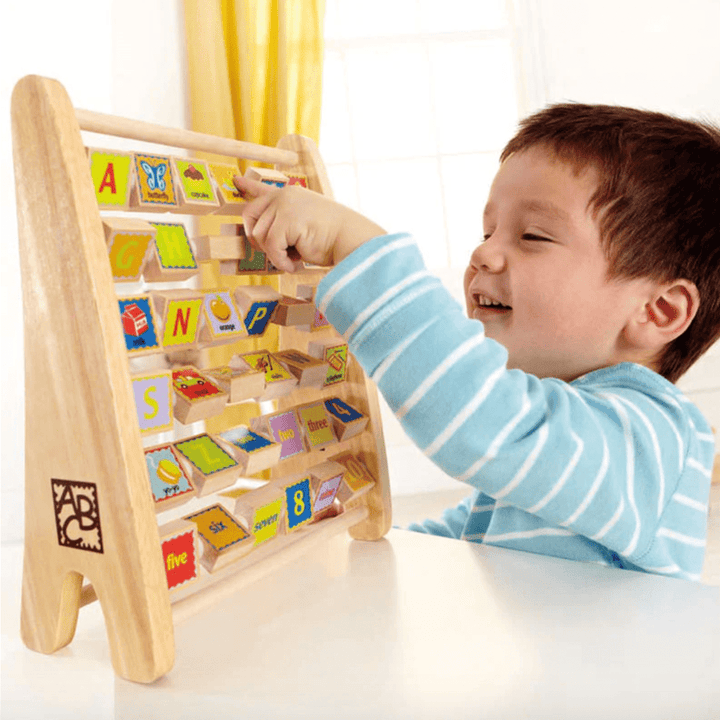 A child interacts with the Hape Alphabet Abacus by Hape, flipping wooden tiles that display letters, numbers, and images on an easel-style frame, enhancing their ABC and numbers knowledge.
