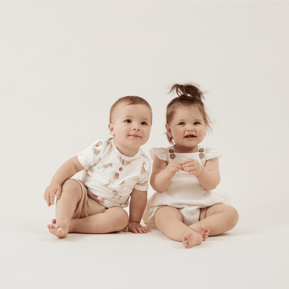 Two toddlers sitting together, one wearing a safari-themed t-shirt with three buttons at the neckline, with taupe shorts, and the other wearing a gingham playsuit