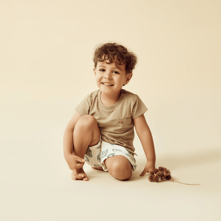 A young child with curly hair, wearing a beige shirt and Wilson & Frenchy Organic Cotton Kids Shorts - LUCKY LASTS - 4 & 5 YEARS ONLY, smiles while crouching on the floor, holding dried flowers.