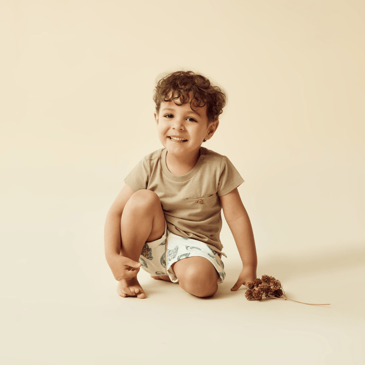 A young child with curly hair, dressed in a beige shirt and Wilson & Frenchy Organic Cotton Kids Shorts - LUCKY LAST - PETIT PUFFIN - 4 YEARS ONLY, sits on the floor smiling while holding dried flowers against a plain beige background.