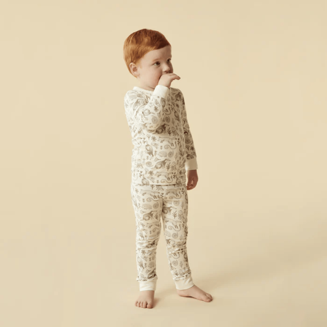 A young child with red hair, in Wilson & Frenchy Organic Long Sleeved Pyjamas with a floral pattern, stands on a beige background, sucking their thumb.