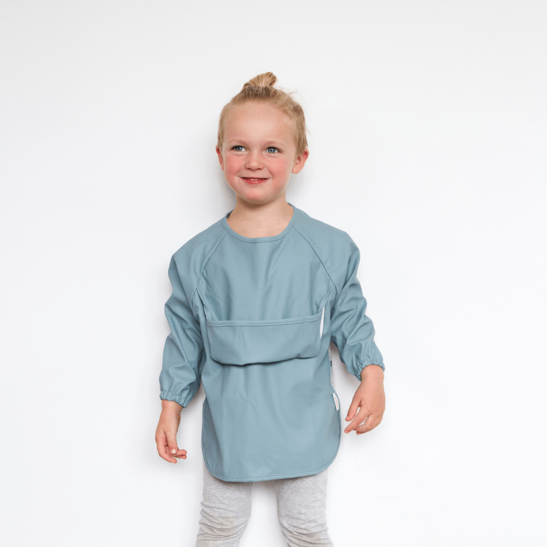 A child wearing a blue Zazi Recycled Full-Sleeved Bib stands against a plain white background, smiling and looking slightly to the side. The waterproof, stain-resistant bib keeps their outfit clean and vibrant.