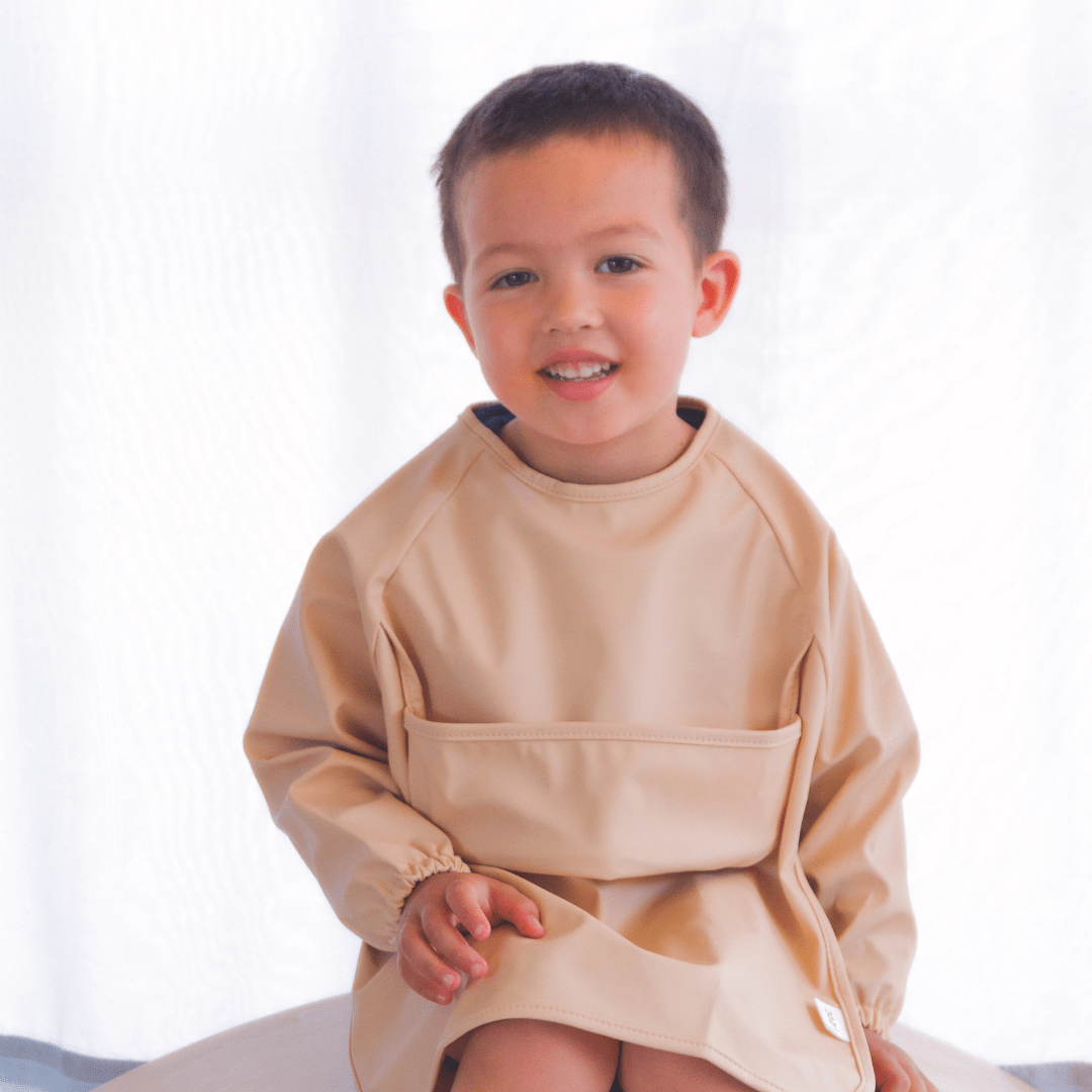 A young child, dressed in a beige smock, sits on a white surface against a white background, sporting the stylish Zazi Recycled Full-Sleeved Bib.