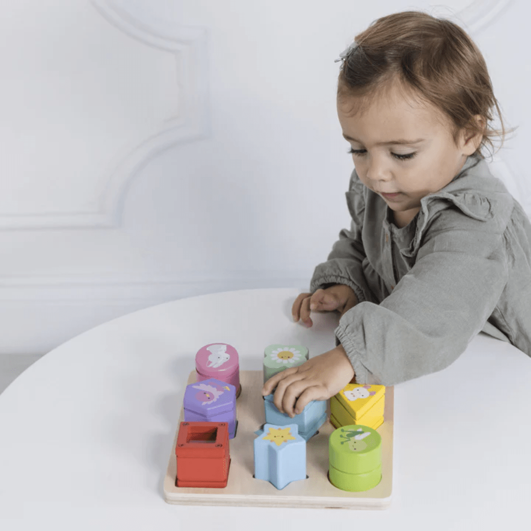 A toddler engages with the Le Toy Van Petilou Sensory Shapes - LUCKY LAST, a colorful wooden shape-sorting toy on a white table, enhancing fine motor skills.