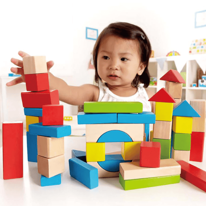 A child is playing with a variety of colorful Hape Build Up and Away Blocks coated in child-safe paints, arranging them into different structures on a table, sparking imaginative play.