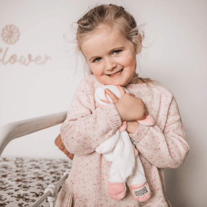 A young girl with light hair, wearing a pink sweater, smiles while holding the Maud N Lil Organic Bunny Comforter - Gift Boxed close to her chest. She stands in a room with a floral-patterned surface and decor in the background.