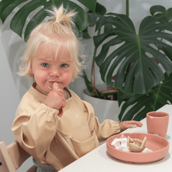 A small child with blonde hair is eating while sitting at a table, wearing a Zazi Recycled Full-Sleeved Bib. The child has food on their face and hands, and there is a pink plate and cup on the table with a plant in the background.