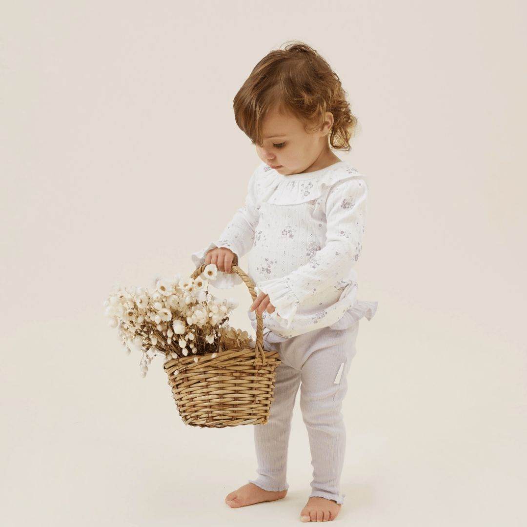 A young child wearing Aster & Oak Organic Rib Lavender Leggings and a white outfit holds a wicker basket filled with dried flowers while standing on a plain background.