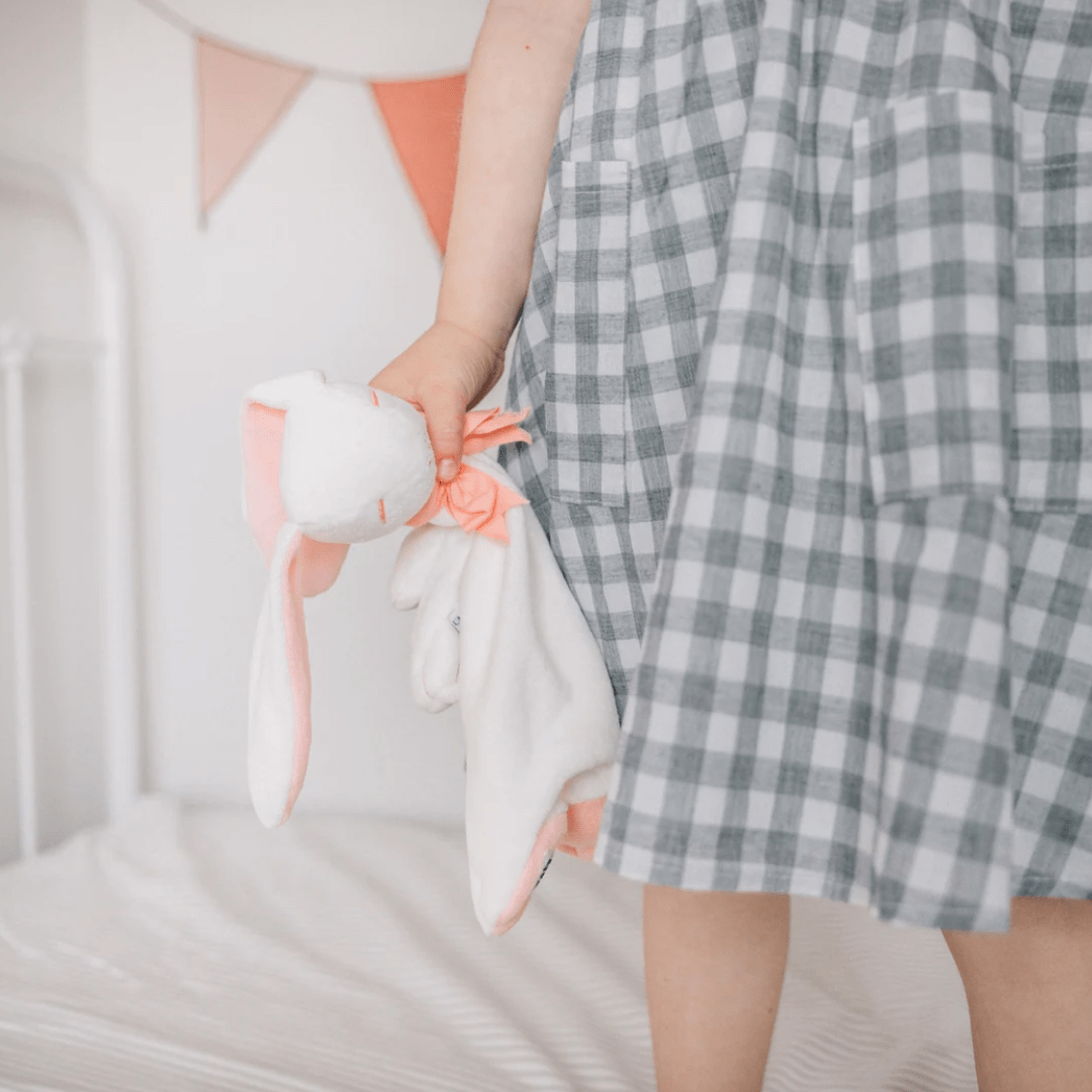 A child wearing a grey and white checkered dress holds a Maud N Lil Organic Bunny Comforter - Gift Boxed by the ear, standing next to a bed with a white bedsheet.