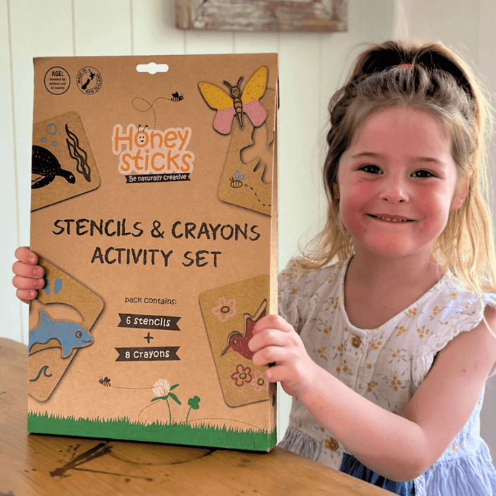 A young girl smiles while holding a Honeysticks Jumbo Stencil & Crayon Activity Set. The eco-friendly box includes 6 stencils and 8 beeswax crayons.