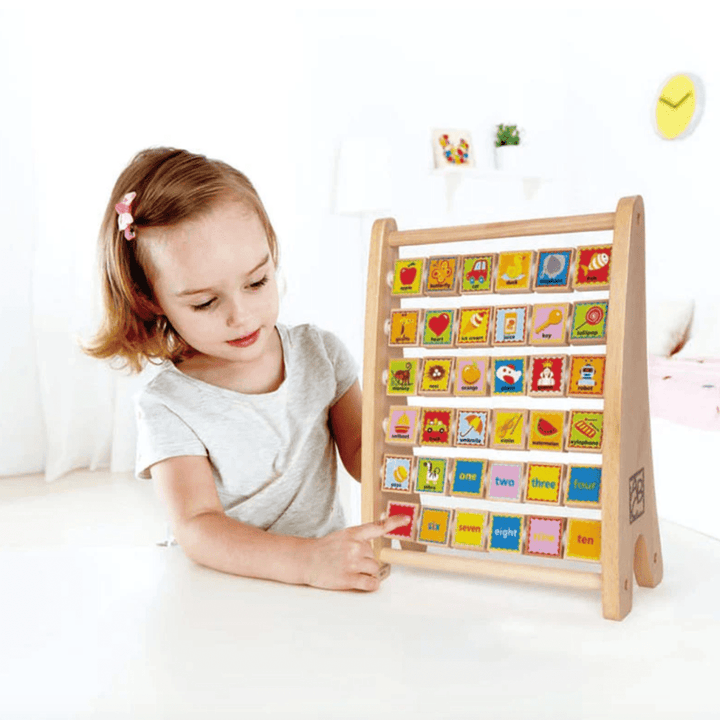 A young girl points at a colorful wooden educational board with ABC and numbers. The board stands on a table in a bright room, showcasing the perfect Hape Alphabet Abacus by Hape.