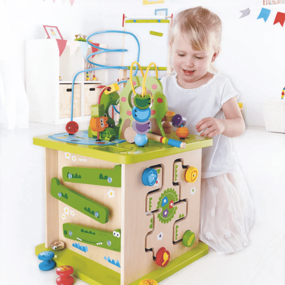 A child in a white dress is on a safari adventure, playing with the Hape Wildlife Safari Adventure Centre. This colorful interactive activity cube is made from durable wood and supports cognitive development in the bright room adorned with cheerful decorations.