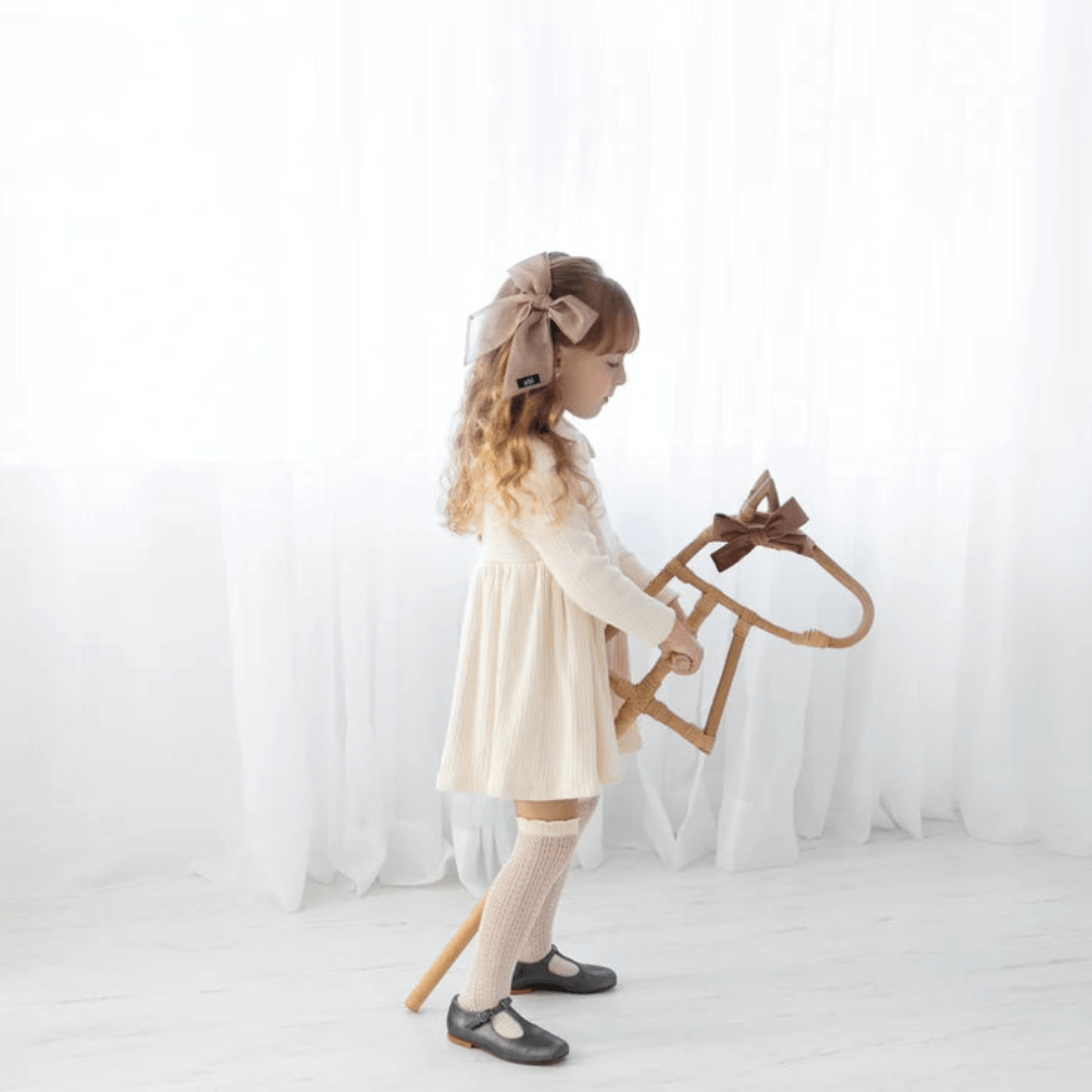 A young girl with a large bow in her hair stands indoors holding the Classical Child Rattan Hobby Horse - LUCKY LAST. She wears a white dress, knee-high socks, and black shoes, engaging in imaginative play with her beloved toy from Classical Child.