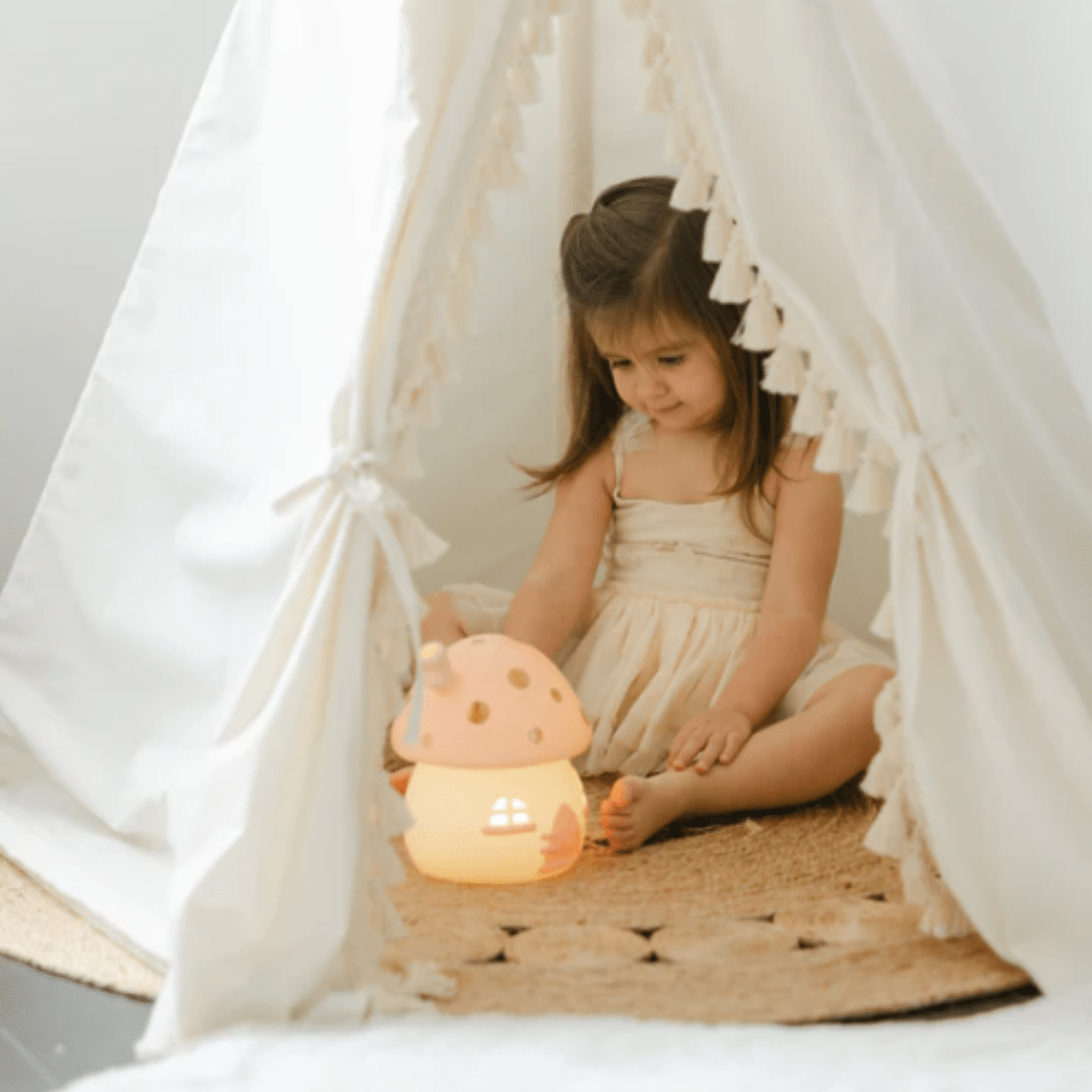 A young girl sits inside a white tent, enchanted by the whimsical charm of a Little Belle Nightlights Ceramic Nightlight - LUCKY LASTS - FAIRY HOUSE ONLY, its gentle glow illuminating the intricate details of the handcrafted porcelain mushroom-shaped lamp on the floor.