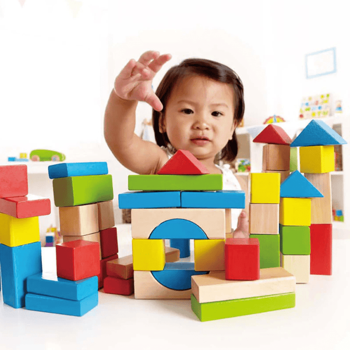A young child with short dark hair reaches out over a colorful assortment of Hape Build Up and Away Blocks, coated in child-safe paints, some stacked and others in various shapes, on a table in a brightly lit room, setting the stage for imaginative play.