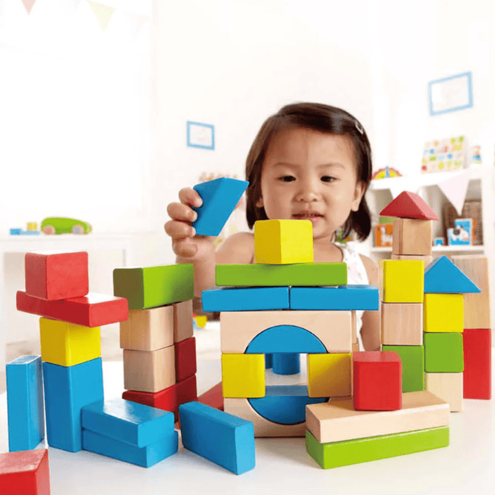 A young child engages in imaginative play with the Hape Build Up and Away Blocks from Hape, stacking them to create various structures on a table in a bright, well-lit room. The child-safe paints ensure a worry-free experience for the parents.