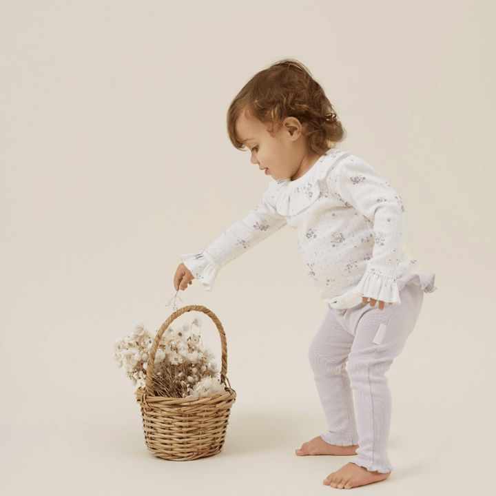 A toddler with curly hair, wearing a white long-sleeved top and Aster & Oak Organic Rib Lavender Leggings, bends to a basket filled with fluffy white items placed on a beige background.