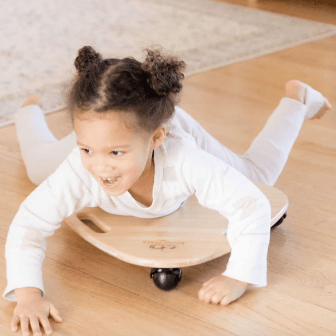 A child wearing a white outfit is lying stomach-down on a Kinderfeets Tummy Glider with wheels, smiling and playing on a wooden floor, enhancing their gross motor skills.