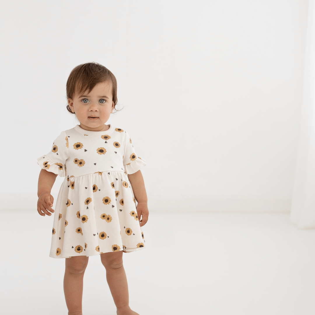 Brown-haired toddler girl wearing an off-white dress featuring bright-yellow sunflowers all-over, ruffle sleeves, and a full skirt