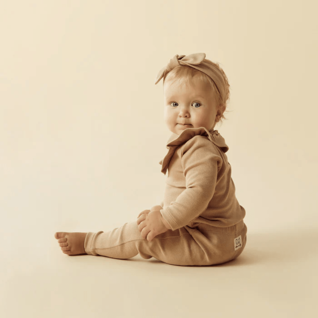 A baby dressed in a beige Wilson & Frenchy Organic Rib Ruffle Top and a matching headband sits on a beige background, looking towards the camera.