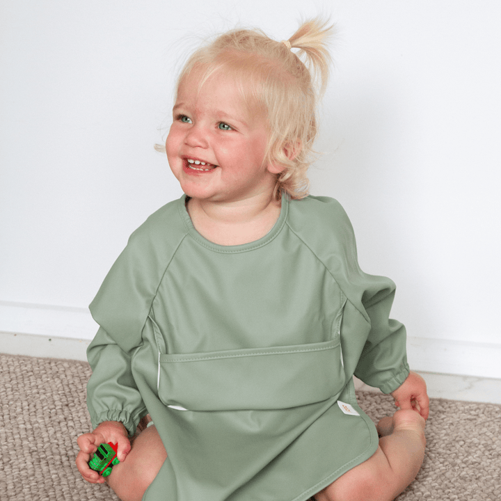 A toddler with blonde hair, wearing a light green outfit, sits on the floor while holding a small green toy in their hand, protected by our Zazi Recycled Full-Sleeved Bib.