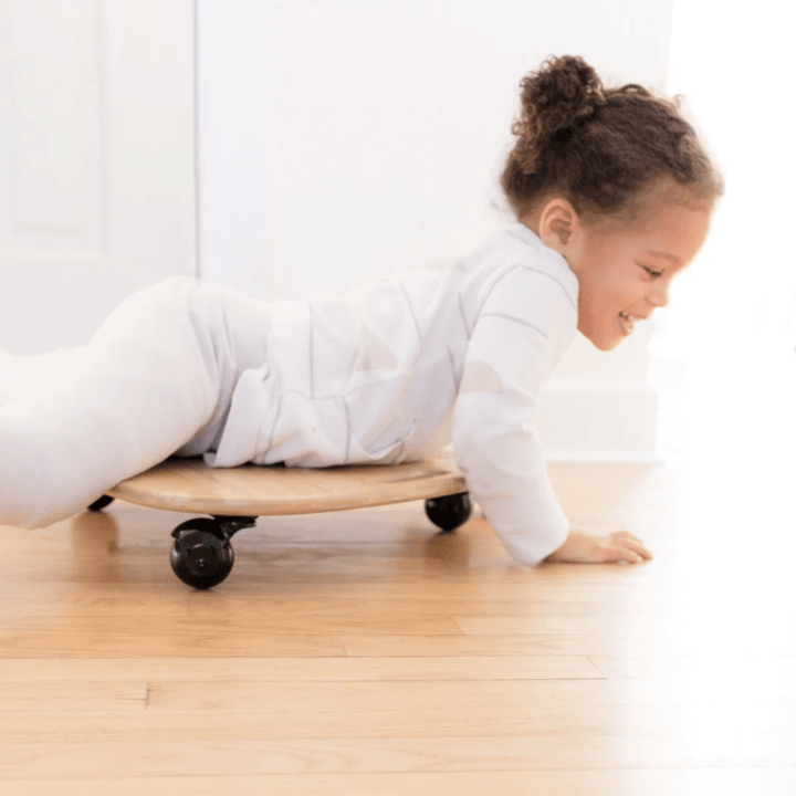 A child lying on a Kinderfeets Tummy Glider with caster wheels, smiling and pushing themselves forward on a wooden floor, enhancing their gross motor skills.