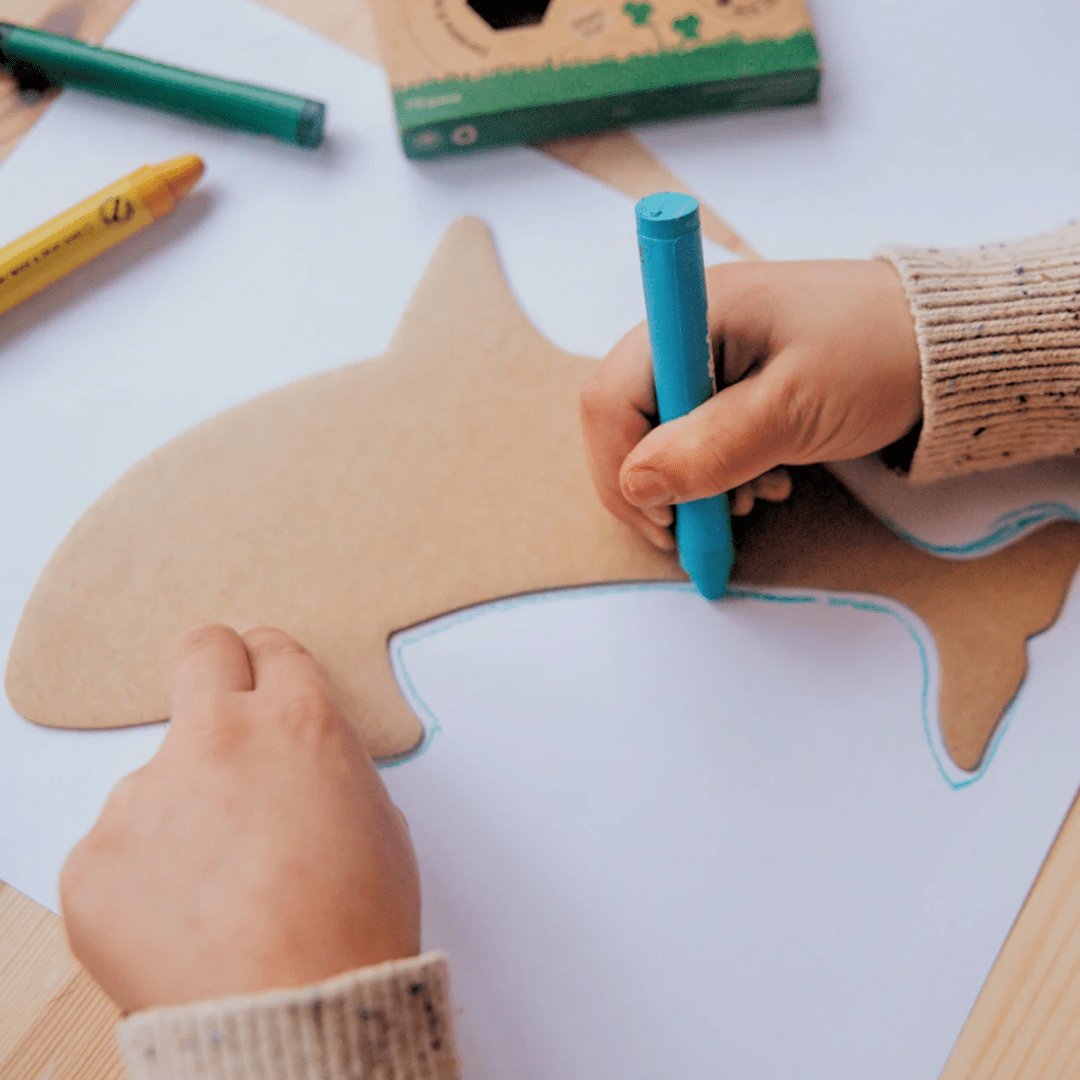 A person traces the outline of a cardboard shark with a blue marker on a piece of white paper. Nearby are other markers, the Honeysticks Jumbo Stencil & Crayon Activity Set, and a green cardboard box from Honeysticks.