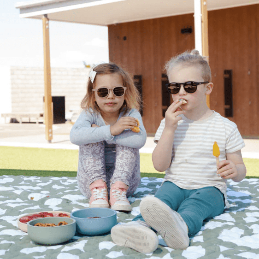 Two children sitting on a Zazi blanket enjoying their food on a summer day.