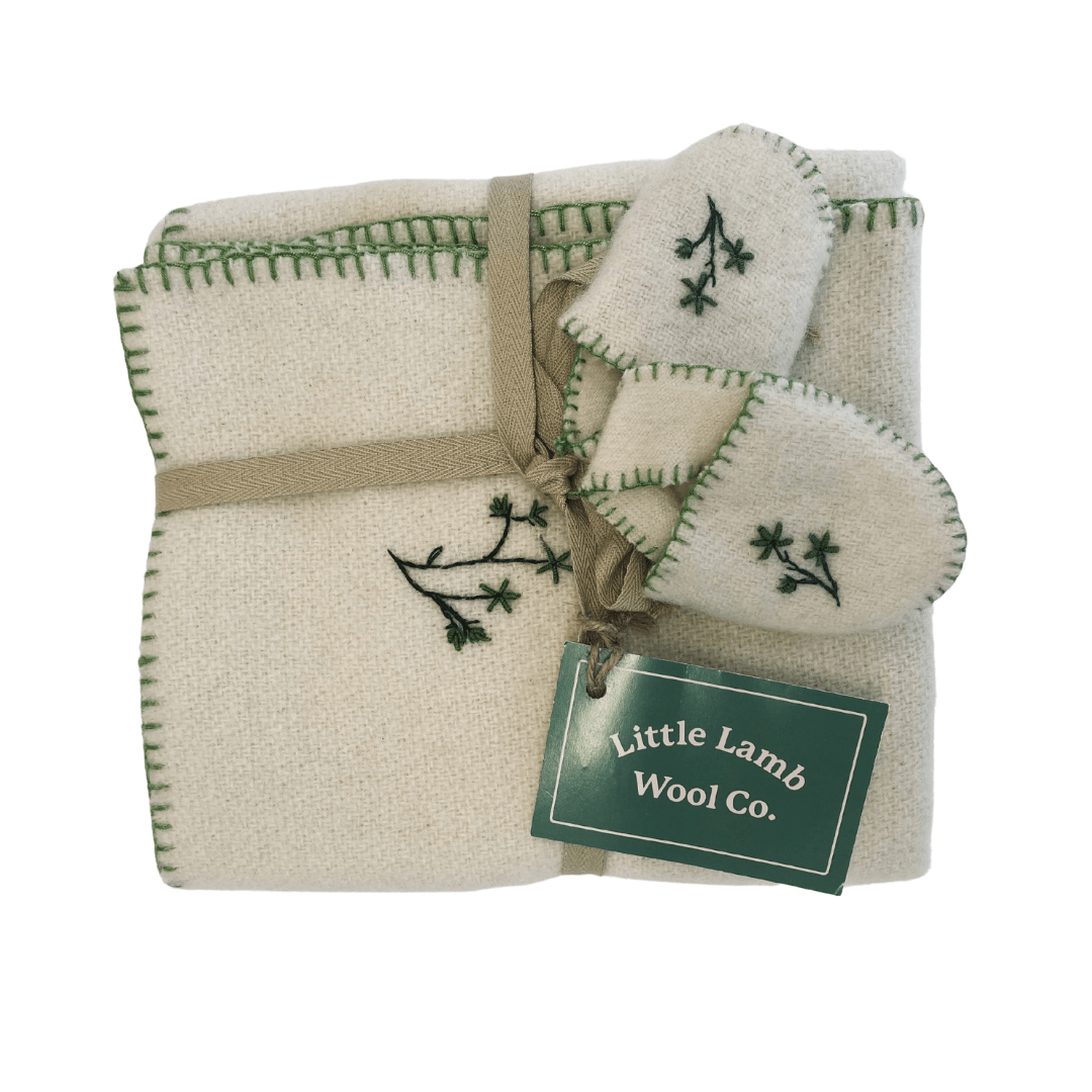 This heirloom-quality upcycled wool blanket from Little Lamb Wool Co., featuring green stitching and floral embroidery, is tied with a brown ribbon. It comes with matching wool booties. A tag reads "Little Lamb Wool Co.", promising timeless charm and cozy warmth for generations to come.