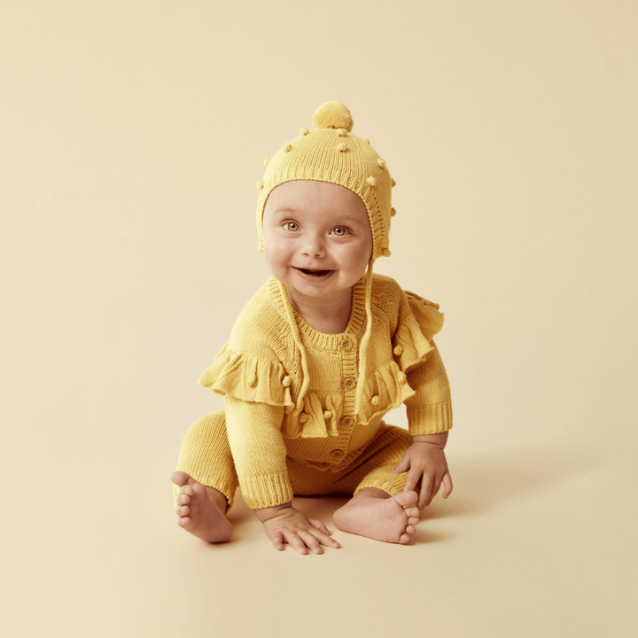 A smiling baby dressed in a yellow knitted outfit with a Wilson & Frenchy Knitted Bauble Bonnet sits against a beige background.