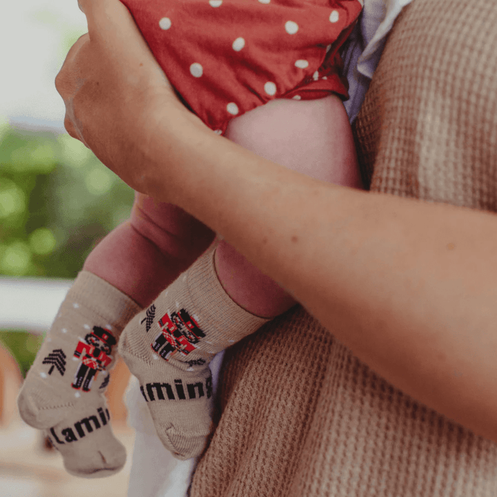 This adorable final sale item showcases a woman lovingly cradling a baby while both are adorably decked out in a pair of Lamington Merino Wool Christmas Socks - Crew - Nutcracker - LUCKY LASTS - 8-12 YEARS ONLY.