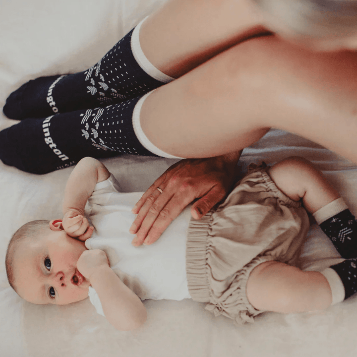 A woman with a baby laying on a bed with Lamington Merino Wool Christmas Socks - Crew - Comet - LUCKY LAST - 4-6 YEARS on.