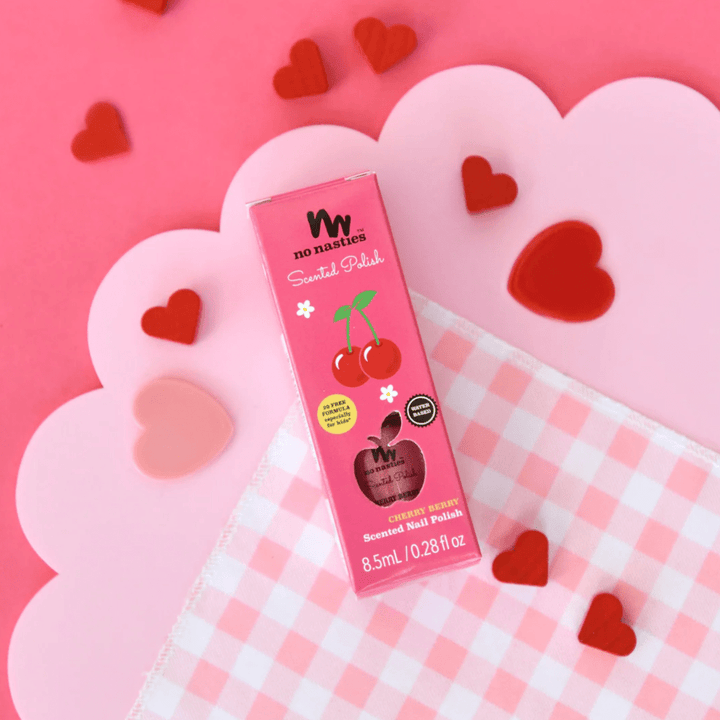 A bottle of No Nasties 20 FREE Kids Scented Nail Polish in Cherry Berry scent sits on a checkered cloth, surrounded by small red and pink heart decorations on a pink background. This non-toxic nail polish from No Nasties is perfect for kids.