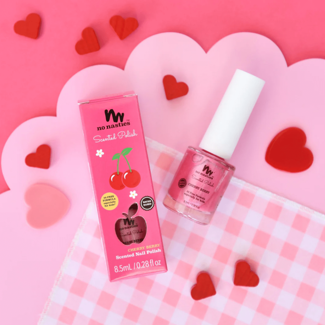 A bottle of No Nasties 20 FREE Kids Scented Nail Polish in pink, labeled "Cherry Berry," sits next to its matching box, surrounded by red heart decorations on a pink and white checkered cloth.