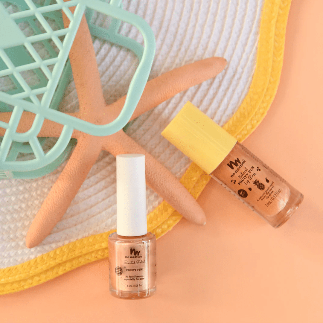 Two bottles of No Nasties 20 FREE Kids Scented Nail Polish, accompanied by beach-themed props such as a starfish, a green basket, and a white sun hat on a yellow-trimmed mat.